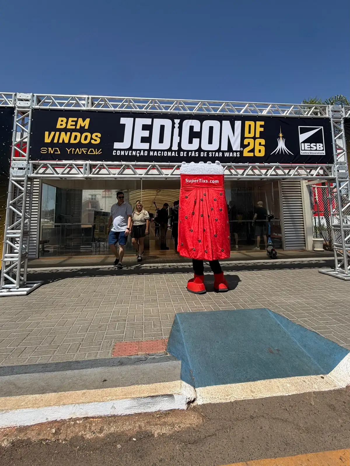 Pórtico de entrada da Jedicon DF 26 com faixa de boas-vindas e o mascote Ticão em frente à porta